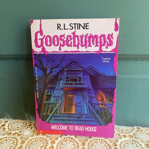 May include: A paperback book titled Goosebumps Welcome to Dead House by R.L. Stine. The cover features a dark blue, eerie house at night with a large tree, framed by a bright pink border with dripping effects and the text It will just kill you.