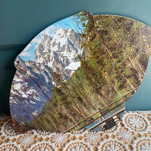 May include: A vintage paper fan featuring a scenic mountain landscape with snow-capped peaks, lush green trees, and a wooden fence. The fan has a curved shape and is made of layered paper with a hole at the top.