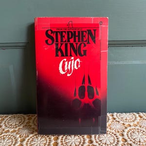May include: A paperback book titled "Cujo" by Stephen King. The cover is predominantly red with black text and a paw print design. The book is set against a green background.