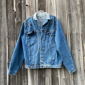 May include: A light blue denim jacket with a light blue collar and embellished with colorful jewels and gold-colored studs. The jacket has a button-up front, two chest pockets, and long sleeves. The jacket is hanging on a clear hanger.