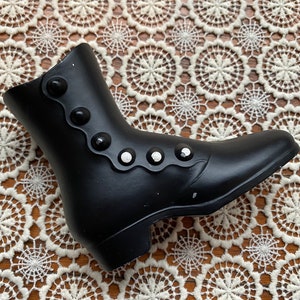 May include: A black decorative boot with a low heel and a row of decorative buttons. The boot is made of a smooth material and is set against a lace doily.