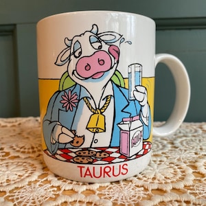 May include: White ceramic mug featuring a cartoon cow dressed in a blue blazer, holding a glass of milk and a cookie. The mug has a yellow background and a red and white checkered pattern at the base, with the word "TAURUS" in red.