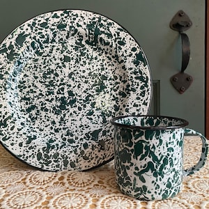 May include: A white and green speckled enamel dinner plate and matching mug. The plate is larger than the mug and both have a black rim.