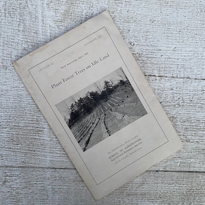 May include: Black and white photo of a pamphlet titled "Plant Forest Trees on Idle Land" published by the Pennsylvania State College School of Agriculture. The pamphlet is on a white wooden surface.