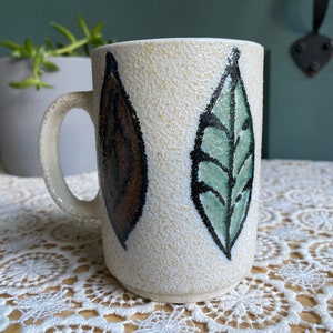 May include: Cream-colored ceramic mug with a textured surface. The mug features two stylized leaf designs, one brown and one green, outlined in black. The handle is curved and matches the mug's color. A decorative item for the kitchen.
