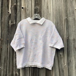 May include: A short-sleeved polo shirt with a textured, bubble-like pattern in pastel colors of pink, blue, and white. The shirt has a white collar and cuffs, and is hanging on a clear hanger against a weathered wooden background.