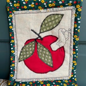 May include: A red apple with green leaves and a small white bird stitched on a beige fabric background. The fabric is framed with a green fabric with a white and yellow floral pattern.