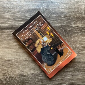 May include: A book titled "Cursing and Other Stories" by Vivian Vande Velde. The cover features an illustration of a woman in a dark dress and a white shawl, standing in a room with a computer and candles. A black cat is sitting on the floor.