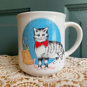 May include: A white ceramic mug featuring a graphic illustration of a grey and white tabby cat wearing a bright red bow tie. The cat is centered within a blue oval frame, accompanied by a yellow bell with green holly leaves and a pink ribbon.