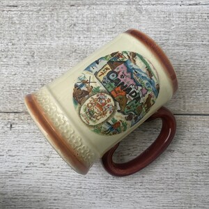 May include: A ceramic mug with a brown handle and a white body. The mug features a colorful illustration of a map of Canada with the word "CANADA" written in a curved arc around the map. The map includes various images of Canadian landmarks and animals.