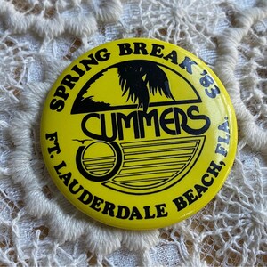 May include: Yellow round button with black text that reads "Spring Break '83 Summers Ft. Lauderdale Beach, Fla.". The button has a black silhouette of a palm tree and a sunset with a wave.