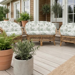 Könnte beinhalten: Gartenmöbel-Set aus Korbgeflecht mit floral gemusterten Kissen. Das Set umfasst eine Doppelsitzbank und zwei Sessel. Die Kissen haben einen weißen Hintergrund mit grünen botanischen Mustern. Topfpflanzen und Bäume sind im Hintergrund zu sehen.