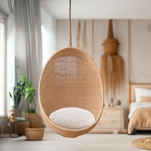 Puede incluir: Una silla colgante de mimbre en forma de huevo con un cojín blanco. La silla está suspendida del techo por una cuerda y se encuentra en una habitación con una cama en el fondo.