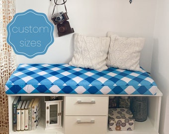 May include: A white bench with blue and white checked upholstery. The bench has two white drawers and a shelf below. The bench is decorated with two white cushions, a vintage camera, and a clock.