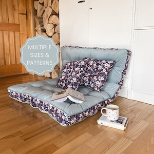 May include: A light blue floor cushion with a floral patterned fabric. The cushion is folded into a chair shape with two floral patterned pillows on top. The cushion is sitting on a wooden floor.
