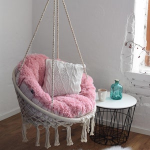 Puede incluir: Una silla colgante rosa y blanca con una almohada de punto blanco. La silla est&aacute; suspendida del techo con una cuerda y tiene una franja alrededor de la parte inferior.