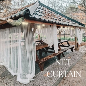 Puede incluir: Un cenador de madera con techo de paja y cortinas blancas transparentes. El cenador tiene una mesa de madera y bancos en el interior. El texto "NET CURTAIN" está escrito en el suelo delante del cenador.