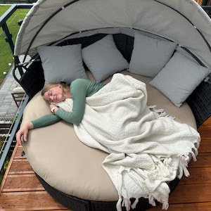 Puede incluir: Una cama redonda al aire libre con dosel, con un cojín beige y cuatro almohadas grises. Una persona descansa en la cama, cubierta con una manta blanca. La cama tiene un marco tejido negro.