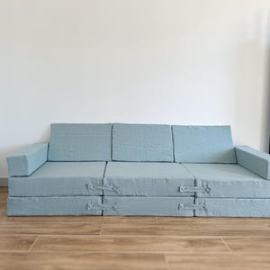 Op de afbeelding: Een lichtblauwe modulaire bank met een eenvoudig ontwerp. De bank is gemaakt van verschillende rechthoekige kussens die in verschillende configuraties kunnen worden gerangschikt. De bank heeft armleuningen aan beide uiteinden en staat op een houten vloer.