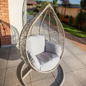 May include: A grey wicker hanging chair with a grey cushion, suspended from a metal stand on a patio.