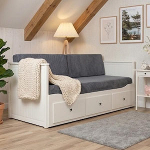 Peut inclure: Un canapé-lit blanc avec un coussin gris et trois tiroirs. Une couverture en tricot crème est drapée sur le lit. Une plante en pot, une petite table d'appoint et des œuvres d'art encadrées complètent l'intérieur cosy.