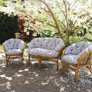Könnte beinhalten: Gartenmöbel-Set aus Korbgeflecht, bestehend aus einer Zweisitzer-Couch und zwei Sesseln. Die Kissen haben ein grau-weißes Muster. Die Möbel stehen auf einer Backstein-Terrasse, mit einem blühenden Baum im Hintergrund.