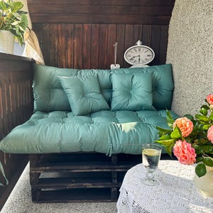 May include: A teal green cushioned pallet sofa with three wooden shelves. The sofa is on a balcony with a white wall and a potted plant. A glass of water and a vase of pink flowers are on a white table with a lace tablecloth.