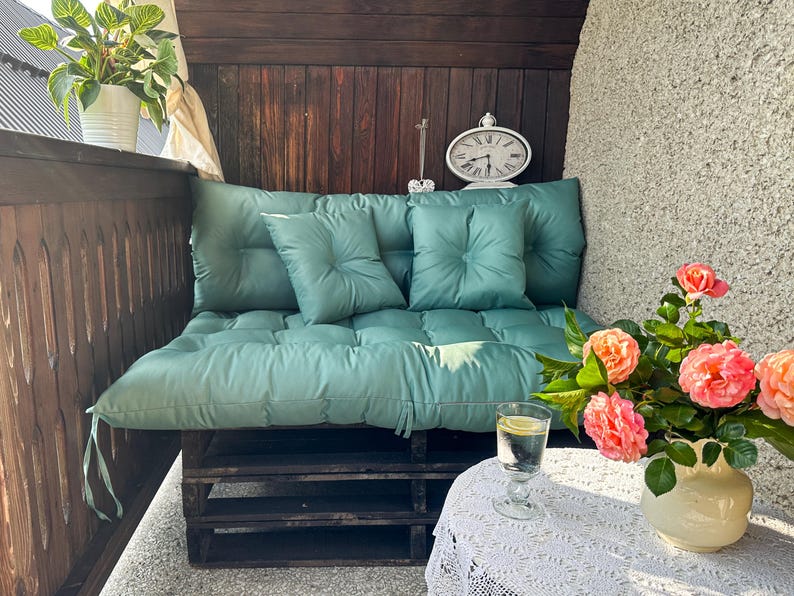 May include: A teal green cushioned pallet sofa with three wooden shelves. The sofa is on a balcony with a white wall and a potted plant. A glass of water and a vase of pink flowers are on a white table with a lace tablecloth.