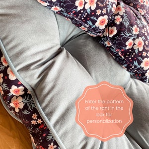 May include: A light blue floor cushion with a floral pattern border. The cushion has a tufted design and is made of soft fabric. A separate floral patterned pillow is on top of the cushion. The text "Enter the pattern of the rant in the box for personalization" is on a pink circle.