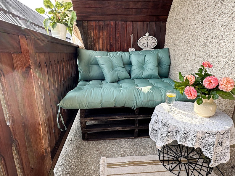 May include: A teal pallet couch with three cushions on a wooden balcony. A white crocheted tablecloth covers a black metal table with a glass of lemonade on top. A bouquet of pink roses in a white vase sits on the table.