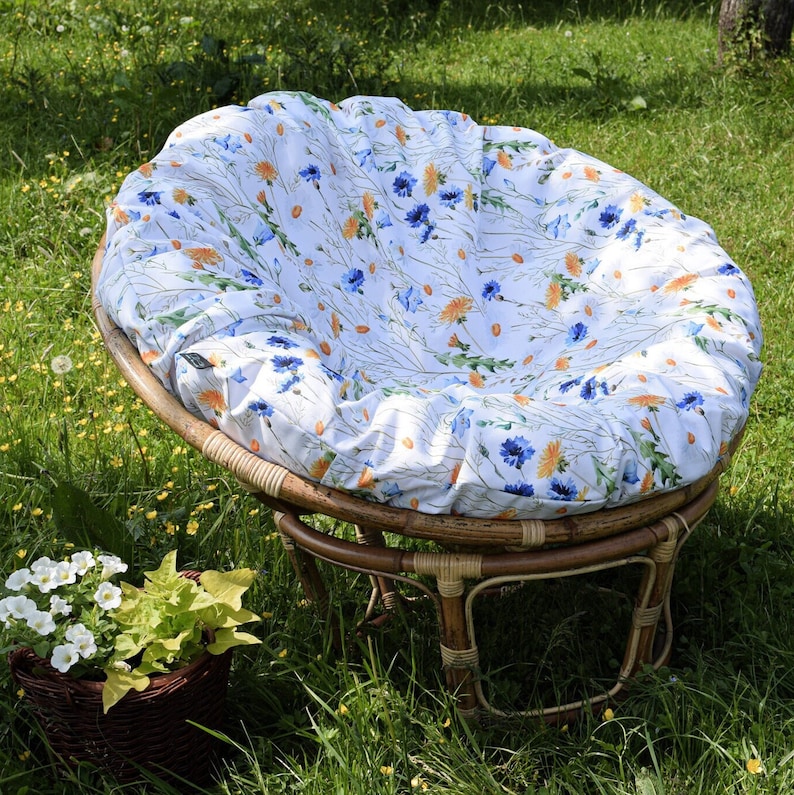 May include: A wicker patio chair with a white cushion featuring a floral pattern of blue and orange flowers. The chair is sitting on a grassy lawn.