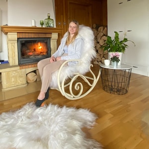 K&ouml;nnte beinhalten: Ein wei&szlig;er Schaukelstuhl aus Kunstfell mit einer Frau, die darin sitzt. Der Stuhl steht vor einem Kamin mit einem Feuer, das darin brennt. Vor dem Stuhl liegt ein wei&szlig;er Kunstfellteppich auf dem Boden.