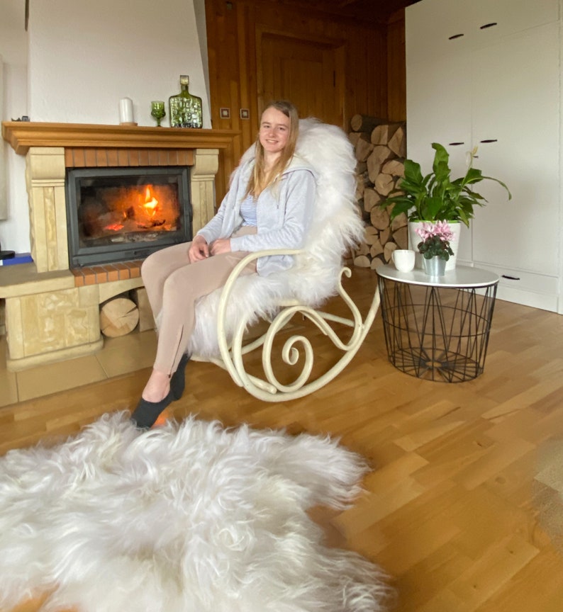 K&ouml;nnte beinhalten: Ein wei&szlig;er Schaukelstuhl aus Kunstfell mit einer Frau, die darin sitzt. Der Stuhl steht vor einem Kamin mit einem Feuer, das darin brennt. Vor dem Stuhl liegt ein wei&szlig;er Kunstfellteppich auf dem Boden.