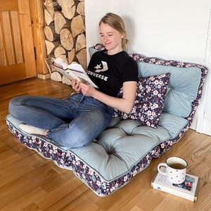 May include: A person sits on a light blue and floral patterned floor cushion with a floral patterned pillow. The person is reading a book and wearing a black t-shirt with the word "Converse" on it and blue jeans.