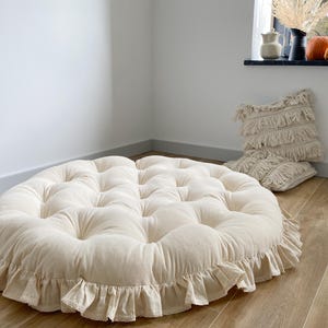 May include: A large, round, tufted floor cushion in a cream color with a ruffled edge. The cushion is made of a soft fabric and has a button-tufted design. The cushion is on a wooden floor.