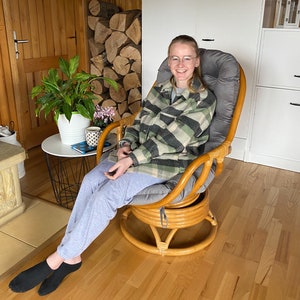 May include: A woman wearing a plaid shirt and grey trousers sits in a brown wicker swivel chair with a grey cushion. The chair is in a room with wood floors and a wooden door in the background.
