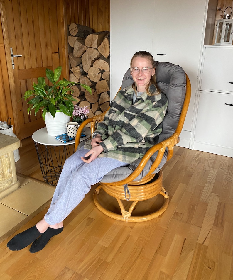 May include: A woman wearing a plaid shirt and grey trousers sits in a brown wicker swivel chair with a grey cushion. The chair is in a room with wood floors and a wooden door in the background.