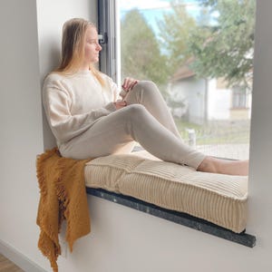 May include: A cream-colored, ribbed cushion sits on a windowsill, with a mustard-colored fringed throw blanket draped over it. A person wearing a cream-colored sweater and pants is seated on the cushion, looking out the window.