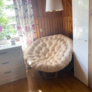 May include: A large, round, cream-colored beanbag chair with a tufted design. The chair is sitting on a wooden floor in a room with a white door and a window with curtains.