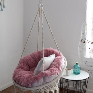 Puede incluir: Una silla colgante de felpa rosa con una almohada blanca, suspendida del techo con una cadena y una cuerda. La silla est&aacute; colgando sobre una alfombra blanca y una mesa auxiliar de metal negro con un jarr&oacute;n de vidrio transparente y una taza blanca.