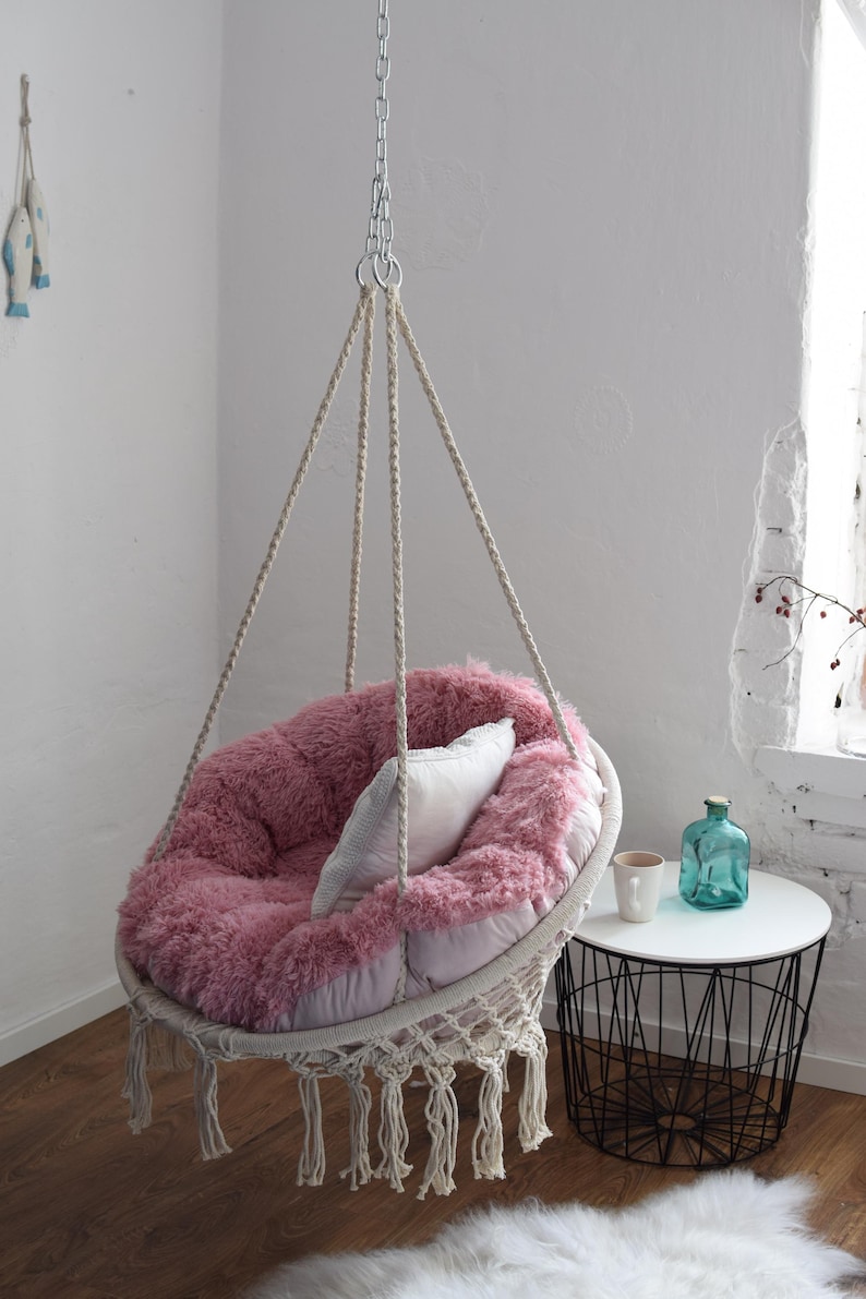 Puede incluir: Una silla colgante de felpa rosa con una almohada blanca, suspendida del techo con una cadena y una cuerda. La silla est&aacute; colgando sobre una alfombra blanca y una mesa auxiliar de metal negro con un jarr&oacute;n de vidrio transparente y una taza blanca.