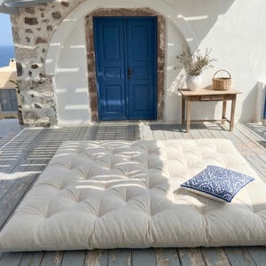 May include: A large, off-white tufted mattress sits on a wooden deck, with a blue and white patterned pillow. A blue door and a small wooden table with a basket and vase are in the background. The scene is outdoors with a view of the sea.