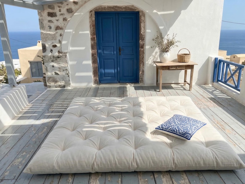 May include: A large, off-white tufted mattress sits on a wooden deck, with a blue and white patterned pillow. A blue door and a small wooden table with a basket and vase are in the background. The scene is outdoors with a view of the sea.