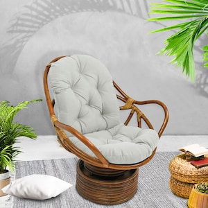 May include: A swivel rattan chair with a light grey cushion. The chair is on a grey rug with a white pillow. The chair is in front of a white wall with a shadow of a palm tree.