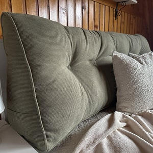 May include: An olive green corduroy headboard with button detailing. Two white, textured throw pillows are on a bed with a cream blanket. A white lamp is visible on the left.