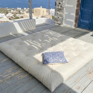 May include: A large, off-white tufted floor cushion with a small blue and white patterned pillow on a weathered wooden deck. The scene is outdoors with a view of the sea and white buildings.