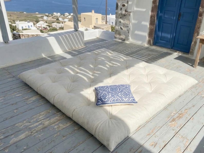 May include: A large, off-white tufted floor cushion with a small blue and white patterned pillow on a weathered wooden deck. The scene is outdoors with a view of the sea and white buildings.