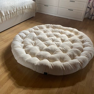 May include: A large, round, off-white cushion with a tufted design. The cushion is made of a soft, plush fabric and has a circular shape.