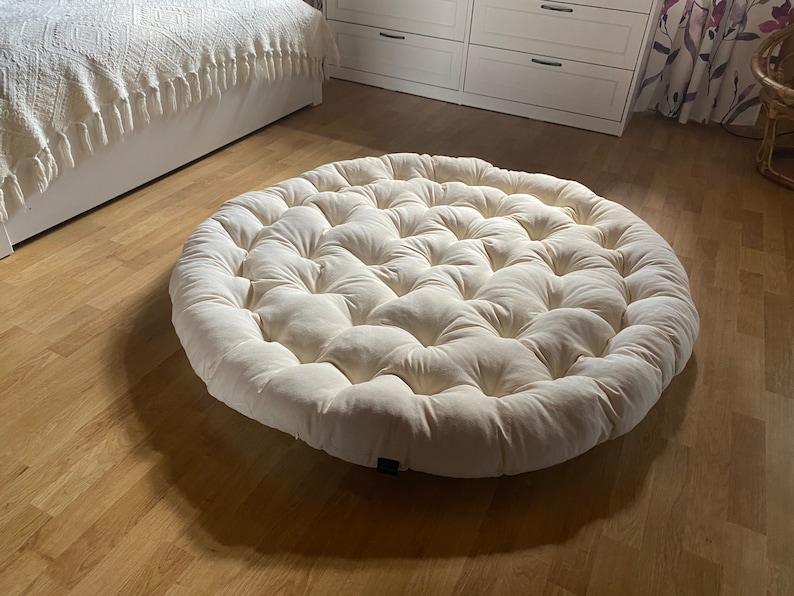 May include: A large, round, off-white cushion with a tufted design. The cushion is made of a soft, plush fabric and has a circular shape.