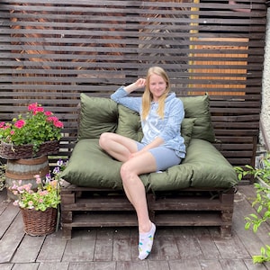May include: A woman sits on a green cushioned pallet couch with a wooden frame. She is wearing a light blue hoodie and grey shorts. The couch is in front of a wooden slat wall.